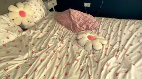 Media: Video of a neatly made bed with a white comforter adorned with small red floral patterns, featuring two large, plush white daisies with pink centers, one in the center and one near the headboard.