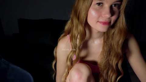 Media: Video of a young Caucasian woman with long, wavy blonde hair and fair skin, wearing a pink tank top, sitting indoors with dim lighting.