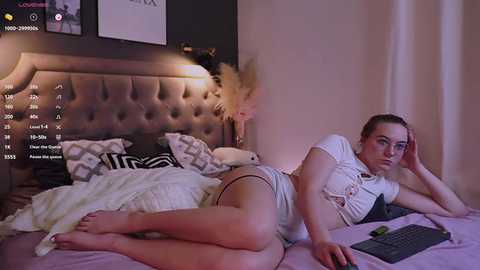 Media: A video of a young woman with light skin and brown hair, lying on a bed with a beige headboard, wearing a white cropped shirt and shorts, holding a laptop, in a cozy, dimly-lit bedroom.