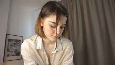 Media: A video of a young woman with fair skin and straight, shoulder-length brown hair, wearing a white button-up shirt, looking down thoughtfully. Background features a beige curtain and a black-and-white framed photo.