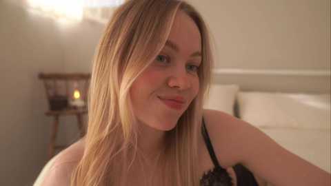 Media: A video of a young woman with fair skin, long blonde hair, and light makeup, wearing a black lace bra, smiling softly in a softly lit bedroom with white bedding and a wooden chair.