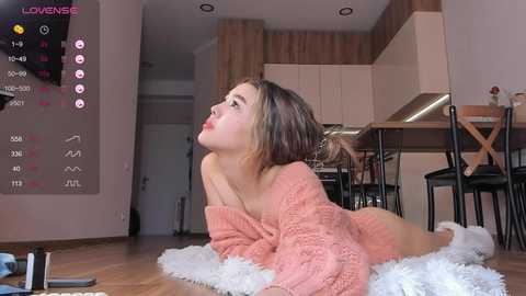 Media: Video of a young, light-skinned woman with brown hair, lying on a white shag rug in a modern kitchen, wearing a peach-colored, off-shoulder sweater.