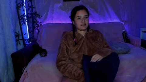 Media: Video of a young woman with dark hair in braids, wearing a brown fur coat and dark pants, sitting on a pink couch in a dimly lit room with purple lighting.