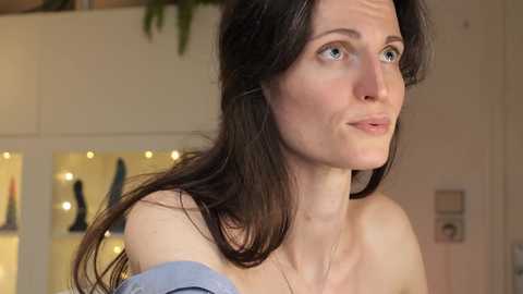 Media: A video of a fair-skinned woman with long dark hair, blue eyes, and freckles, wearing a blue off-shoulder top, looking contemplative indoors.