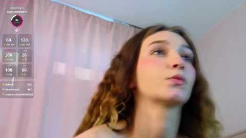 Media: Video of a young Caucasian woman with wavy brown hair and light skin, blushing, looking upward. She's wearing a light-colored top. Background includes a white wall and a pink curtain. Social media app interface on the left displays statistics.