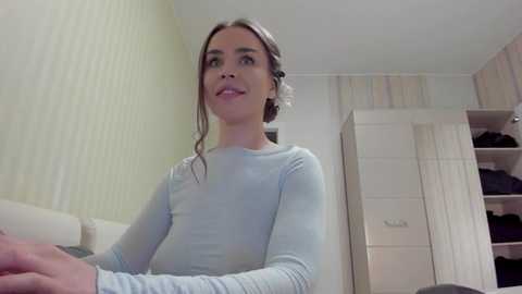 Media: Video of a light-skinned woman with straight brown hair in a bun, wearing a light blue long-sleeved top, standing in a minimalist bedroom with beige wallpaper, white wardrobe, and a bed.