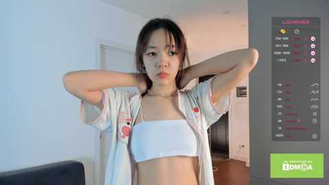 Media: A video of an East Asian woman with long black hair, medium build, and small breasts, wearing a white bra and open floral shirt, posing in a modern, white-walled room with a TV displaying game statistics.