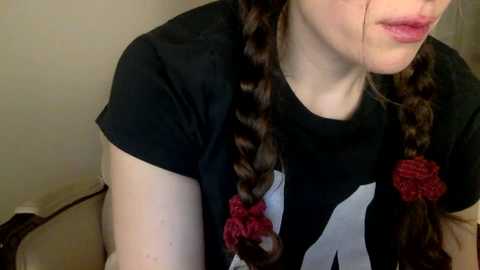 Media: Video of a young woman with fair skin, wearing a black T-shirt with a white graphic, and twin braids adorned with red beads.