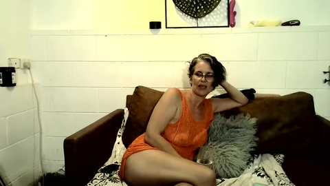 Media: Video of a middle-aged woman with glasses, fair skin, and brown hair, wearing a sleeveless orange dress, lounging on a brown couch in a modestly decorated room with white walls and a fur throw pillow.