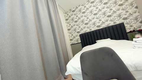 Media: A video of a modern bedroom with a gray upholstered headboard, white bedding, and a patterned wallpaper featuring black and white floral designs. A gray chair and beige curtains are visible.