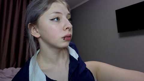 Media: Video of a young, fair-skinned woman with platinum blonde hair, wearing a dark blue blouse, looking contemplative in a dimly lit bedroom with a flat-screen TV on the wall.