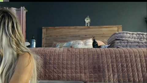 Media: Video of a woman with long blonde hair standing beside a bed with a wooden headboard and pink quilt.
