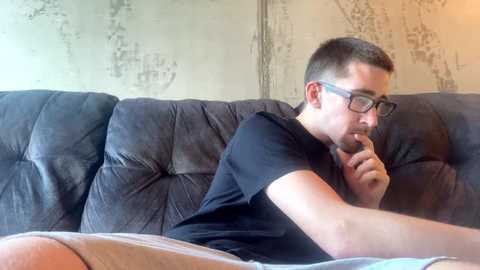 Media: Video of a young man with short hair, glasses, and a beard, resting on a black, tufted couch, holding his finger to his mouth in thought, in a dimly lit room with a textured, gray wall.