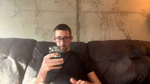 Media: Video of a man with short dark hair, glasses, and black shirt, holding a phone, sitting on a dark, plush couch with a textured fabric, against a textured, beige wall with floral patterns.