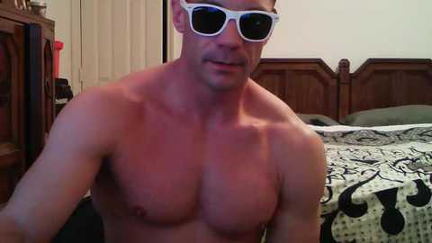 Media: Video of a shirtless, muscular man with short hair, wearing white sunglasses, in a bedroom with a wooden headboard, patterned bedspread, and partially visible furniture.