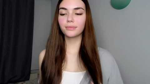 Media: Video of a young woman with long, straight brown hair, wearing a white top, standing against a plain white wall with a dark curtain on the left.