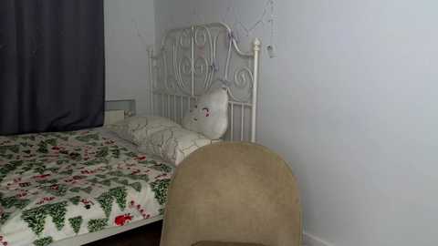 Media: Video of a simple, small bedroom with a white metal bed frame, a beige chair, a gray curtain, and a festive Christmas-patterned duvet.