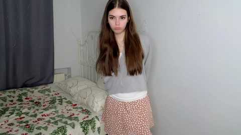 Media: A video of a young woman with long brown hair, wearing a gray sweater and patterned skirt, standing near a bed with floral bedding and gray curtains in a simple, white-walled room.