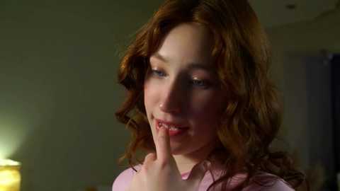 Media: A video of a fair-skinned, red-haired woman with wavy hair, wearing a pink blouse, holding her finger to her lips, in a dimly lit room with a blurred background.