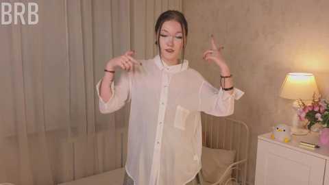 Media: A video of a young woman in a white blouse, standing in a beige room with a bed, lamp, and flowers.