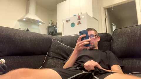 Media: Video of a man in a dark gray sofa, wearing glasses and a black shirt, taking a selfie with a smartphone, kitchen in background.