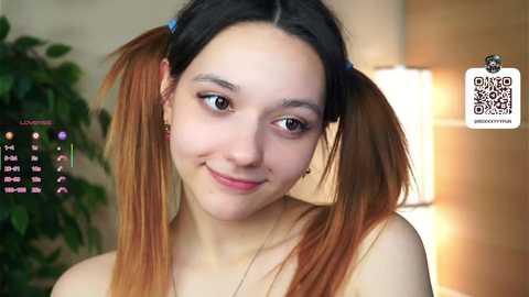 Media: Video of a young Asian woman with light skin, pigtails, and minimal makeup, smiling softly. Background includes a calendar, green plant, and a warm-toned lamp.