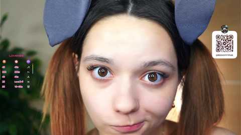 Media: A close-up video of a young Asian woman with light skin and brown eyes, wearing a grey bunny headband. She has long, straight brown hair and is smiling slightly. The background is blurred with green plants and a white wall.