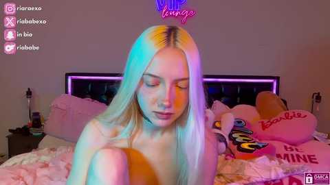 Media: Video of a topless, blonde woman with fair skin and long hair, sitting on a bed in a dimly-lit bedroom, wearing a neon crown, surrounded by plush toys and a \"Be Mine\" sign.