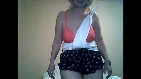 Media: Video of a smiling, light-skinned woman wearing a white seatbelt over a coral bra and black shorts with a floral pattern, against a plain beige wall.