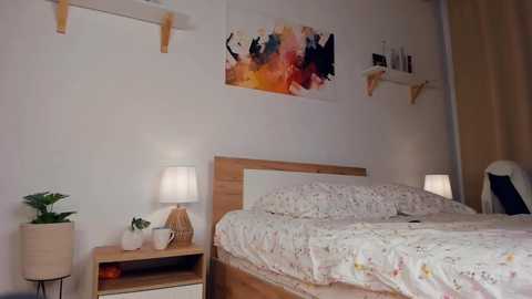 Media: Video of a minimalist bedroom with a wooden bed, white bedding, a wooden nightstand, a lamp, a potted plant, a colorful abstract painting, and white walls with wooden shelves.