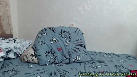 Media: A video of a bed with a blue comforter featuring cartoonish black and white explosions, partially covered by a beige blanket. The room has a plain white wall and a partially visible wooden headboard. The URL \"fansly.com/NovaSasha\" is visible in the bottom right corner.