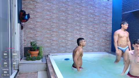 Video of three shirtless, muscular men in swim trunks in a rectangular indoor pool, one wearing sunglasses, set against a brick wall.