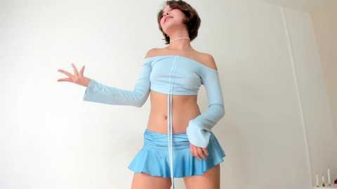 Media: Video of a slim, light-skinned woman with short brown hair, wearing a light blue off-shoulder crop top and matching mini-skirt, against a plain white wall background.