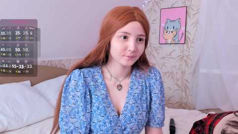 Media: Video of a young woman with long red hair and fair skin, wearing a blue floral dress, sitting on a bed. The room has a calendar overlay, a pink cat poster, and a plaid shirt on the bed.