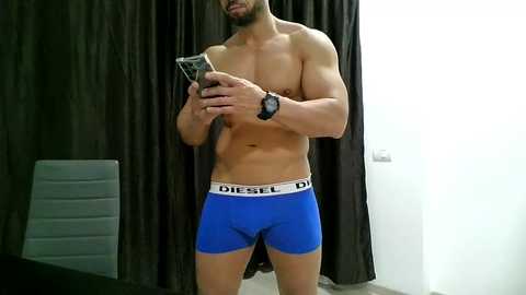 Media: Video of a muscular, shirtless man with light skin tone wearing blue Diesel underwear, holding a smartphone, against dark curtains and a gray bench.