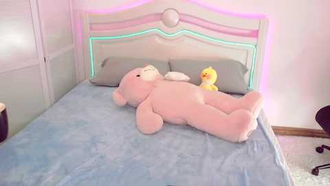 Media: Video of a pink teddy bear lying on a light blue bedspread, surrounded by grey pillows and a yellow duck toy, in a pastel-colored bedroom with a white headboard and pink neon lights.