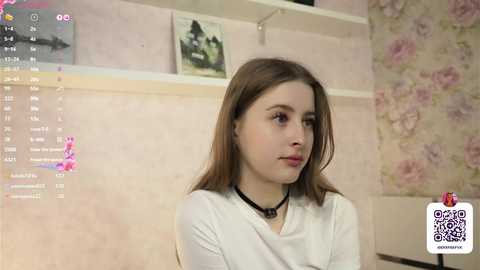 Media: Video of a young Caucasian woman with light skin and straight brown hair, wearing a white top and black choker, in a pink-tiled room with floral wallpaper.