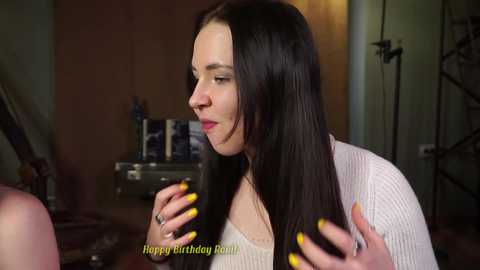 Media: A video of a young woman with long, dark hair, wearing a white, ribbed sweater, and yellow nail polish, smiling. Background includes a bookshelf and a camera tripod.