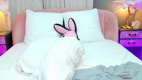 Media: Video of a plush bunny with a pink body, black ears, and white fur, nestled in a neatly made bed with white linens and a pastel pink headboard.