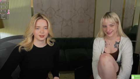 Media: Video of two blonde women with fair skin, one in a black top and the other in a beige cardigan, sitting indoors.