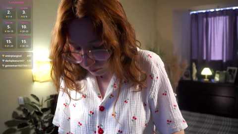 Media: Video of a young woman with red hair, wearing glasses and a floral-patterned shirt, sitting at a desk in a dimly lit bedroom.