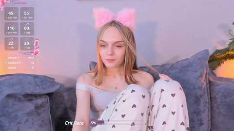 Media: A video of a young Caucasian woman with light skin, straight blonde hair, wearing a pink cat ear headband and white pajamas with heart patterns, sitting on a gray couch.