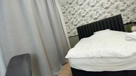 Media: Video of a modern bedroom with a black tufted headboard, white bedding, and a grey upholstered chair. The wallpaper features a botanical pattern in shades of grey.