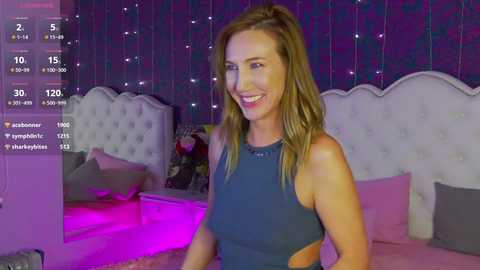 Media: Video of a smiling, fit woman with light skin and long brown hair, wearing a navy blue sports bra, standing in a purple-lit bedroom with a tufted white headboard.