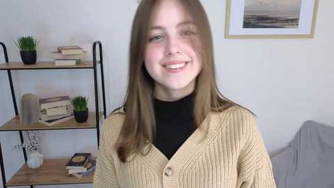 Media: Video of a smiling young woman with straight brown hair, wearing a beige cardigan over a black top, standing in a minimalist room with bookshelves and framed artwork.