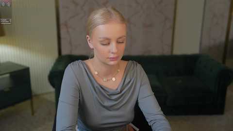 Media: Video of a blonde Caucasian woman with fair skin, wearing a grey long-sleeve top and a gold necklace, sitting indoors, facing a dark green velvet couch, with a beige wall and floral wallpaper in the background.