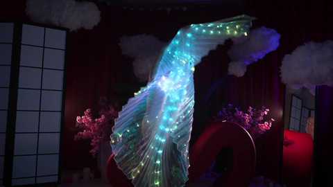 Media: Video of a performer in a colorful, illuminated LED dress, set against dark, red curtains with traditional Japanese sliding doors. The background features pink floral arrangements and smoke effects.