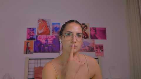 Media: A video of a young woman with fair skin, wearing glasses and a beige top, with a finger on her lips, in a room with pink and purple posters on the wall.