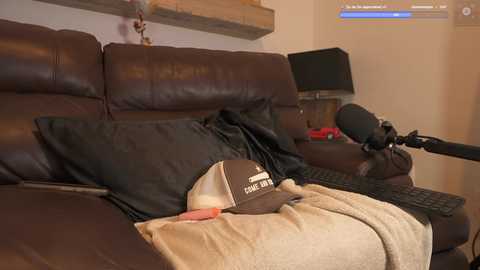 Media: Video of a cozy living room with a brown leather couch, a beige blanket, and a black baseball cap with \"Columbia\" on it. A microphone and computer screen are visible in the background.
