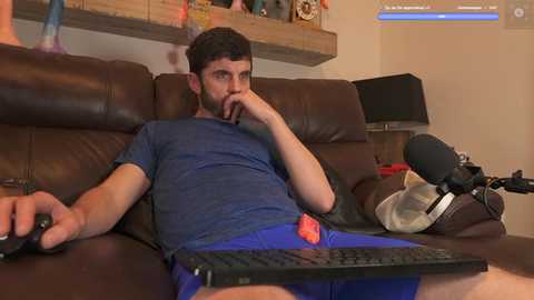Media: A video of a bearded man in a blue shirt and blue shorts, reclining on a brown leather couch, holding a controller, watching a video game on a laptop.